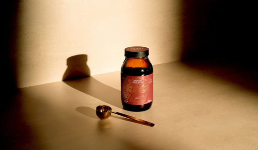 A jar of Radiant Collagyn alongside a golden ritual scoop, in a beam of sunlight on a neutral floor