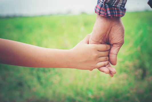 A close up of the hands of a father and child walking together in a green grassy field