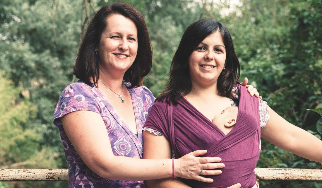 Two women stand together outdoors, one wearing a purple wrap with a baby nestled closely. Lush greenery serves as a backdrop.