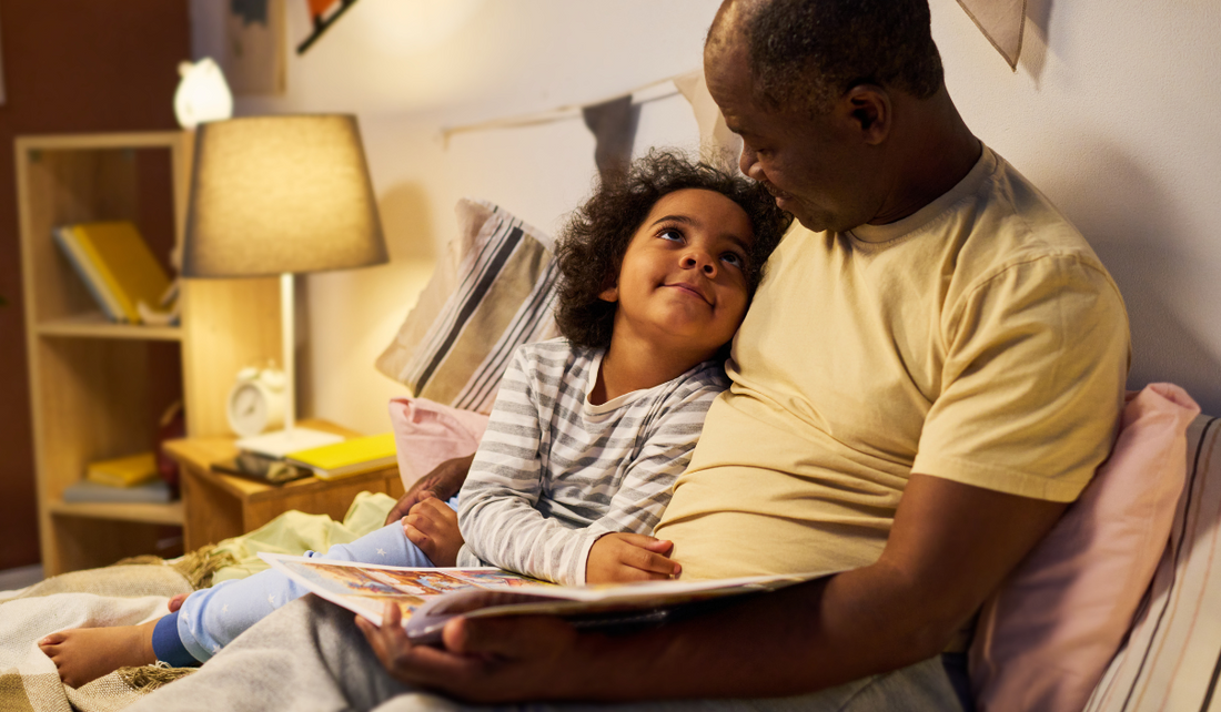 A child snuggles close to an adult while reading a colourful book in a cosy, softly lit bedroom.