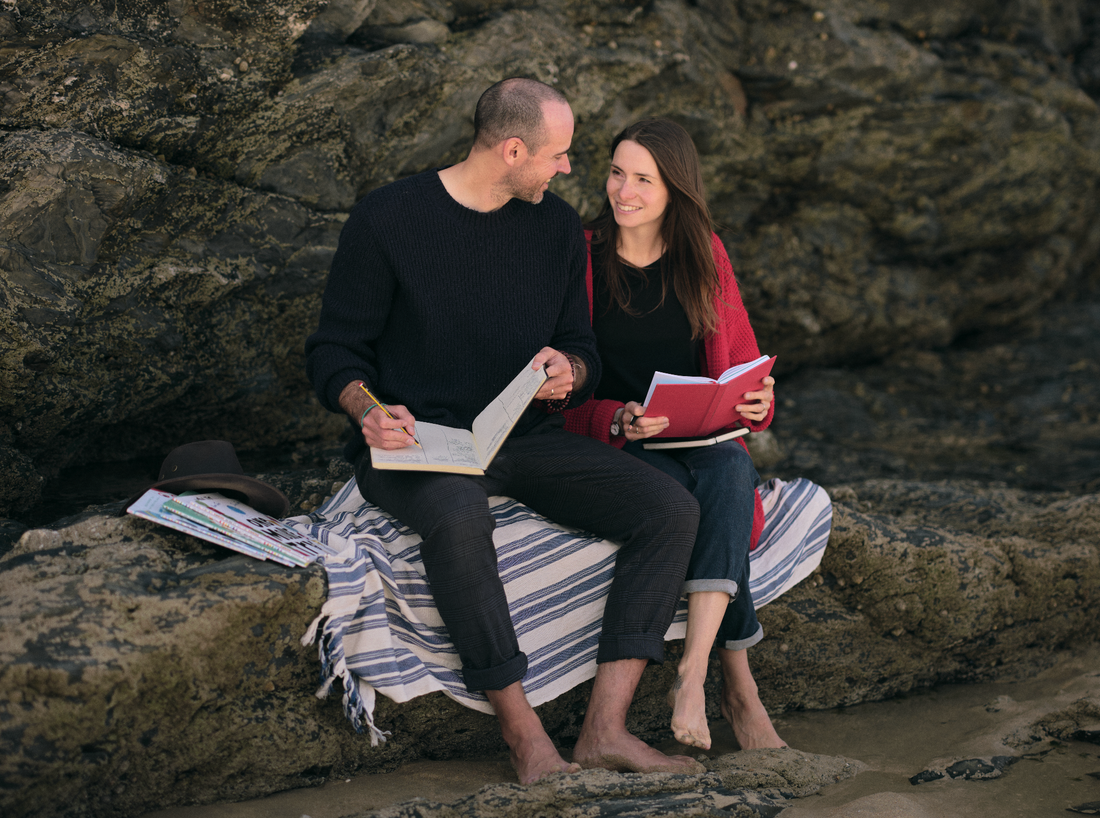 Two people sitting on a striped towel across rocks, one holding a red book and one holding a notebook and pen, smiling
