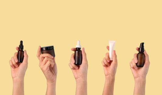 Five hands holding various skin care bottles and tubes against a light beige background.